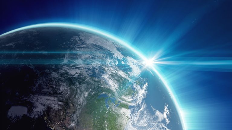 View of Earth from space with the Sun breaking over the horizon, casting a blue glow around the planet. The Earth's surface features clouds, landmasses, and bodies of water. The dark vacuum of space serves as the backdrop.