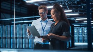 A man and a woman stand in a server room. The man wears glasses and is dressed in business casual attire with a lanyard. The woman smiles and holds an open laptop. Rows of servers with blue lights fill the background, creating a modern and high-tech atmosphere.