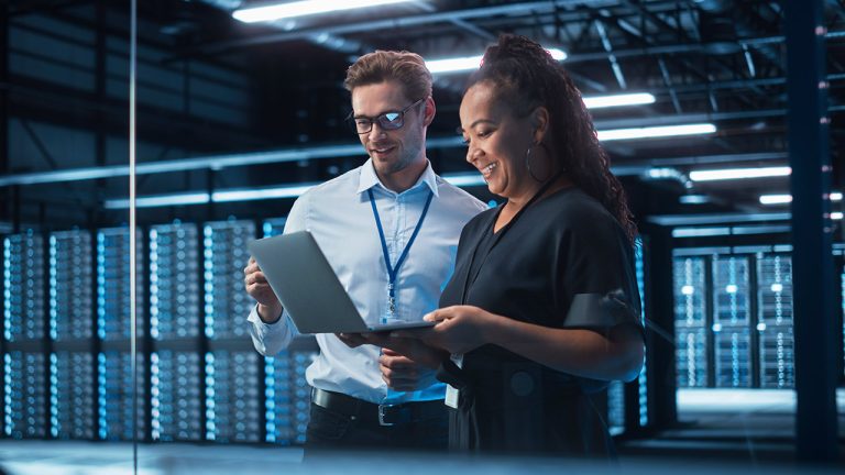 A man and a woman stand in a server room. The man wears glasses and is dressed in business casual attire with a lanyard. The woman smiles and holds an open laptop. Rows of servers with blue lights fill the background, creating a modern and high-tech atmosphere.