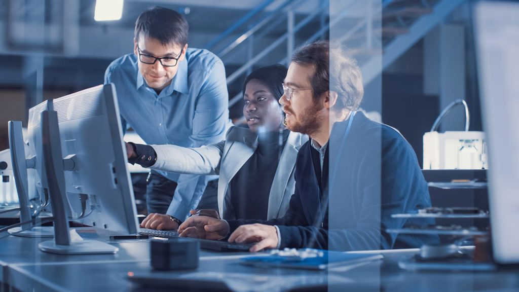 Three people are collaborating in a modern office environment, focusing on a computer screen. One person is standing and pointing at the screen, while the other two sit and type. The scene has a professional and tech-focused atmosphere.