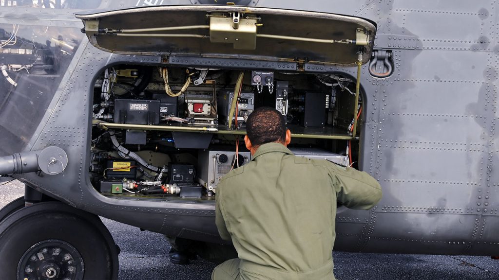 A person wearing a green military uniform is crouching in front of an open maintenance panel on a military aircraft, working on the internal components. The aircraft's grey exterior and various mechanical parts are visible.