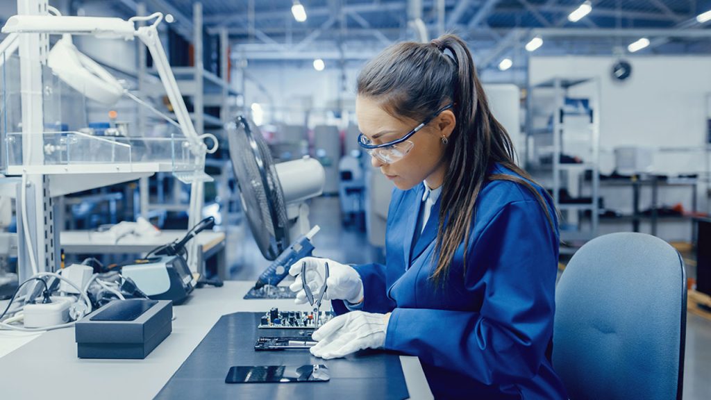 A woman wearing safety glasses and a blue lab coat works on electronic components at a workstation in a modern laboratory. She uses tools for precise adjustments, surrounded by various lab equipment and workstations in the background.