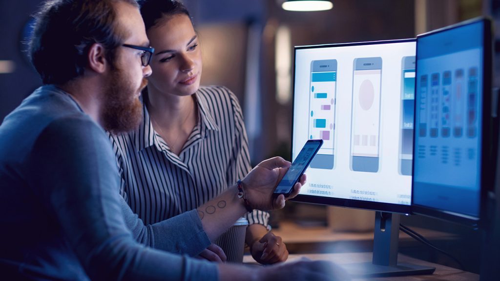 Two professionals, a man and a woman, are collaborating at a desk, looking at a smartphone and a computer screen displaying software or app designs in a dimly lit office.