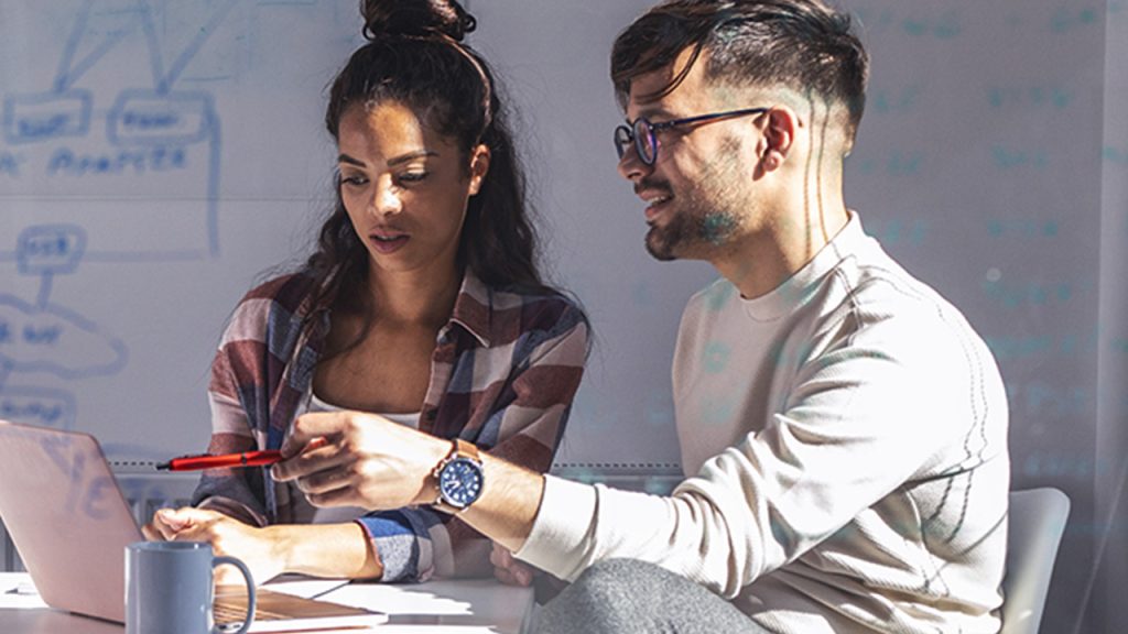 Two professionals, a man and a woman, collaborate over a laptop in a sunny office, with whiteboard markers and coffee on the table, discussing work intensely.