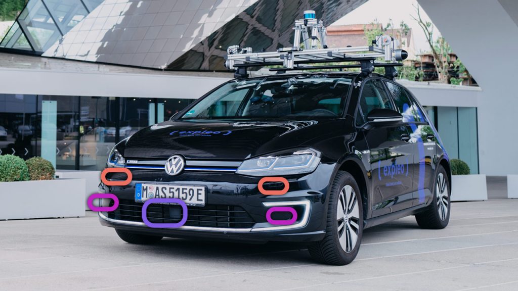 A black Volkswagen car equipped with various sensors and cameras on its roof, parked in front of a modern building. Colored outlines highlight specific sensors on the front of the vehicle.