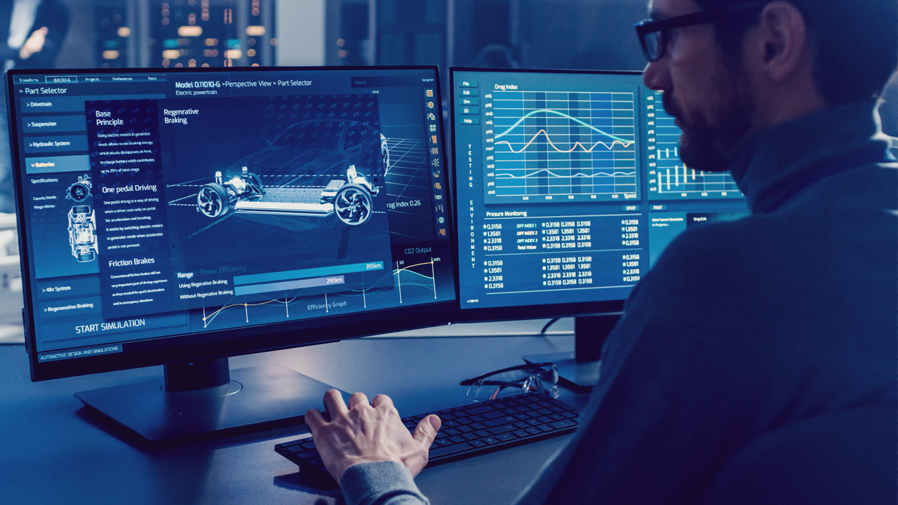 A man at a desk works on a computer with dual monitors displaying technical diagrams, vehicle schematics, data charts, and graphs in a high-tech, modern office setting.