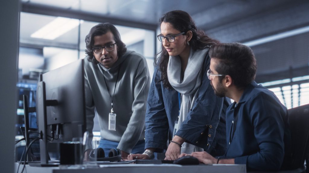 Three people are collaborating in an office environment. One person is seated and working at a computer, while the other two stand nearby, engaging in discussion. The office has a modern, professional atmosphere with glass partitions and various equipment in the background.