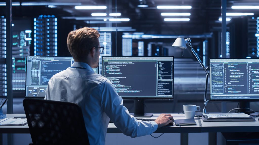 A person with short blond hair wearing glasses is seated at a desk with three computer monitors displaying code. The scene is set in a modern, dimly lit office with rows of server racks in the background. A desk lamp and a cup of coffee are also visible.