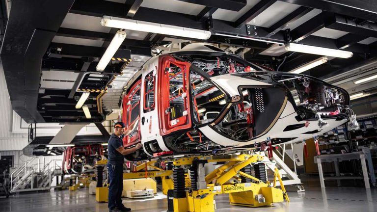 An individual works on the assembly of a helicopter's fuselage inside a factory. The helicopter frame, supported by yellow stands, reveals intricate internal wiring and components. The factory is well-lit with various structural and mechanical elements visible.