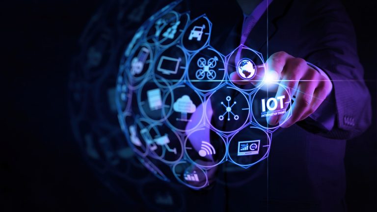 A person wearing a dark suit points at a futuristic, glowing interface displaying various hexagonal icons, including symbols for technology, automation, and communication. The term "IOT" (Internet of Things) is prominently highlighted in the interface.