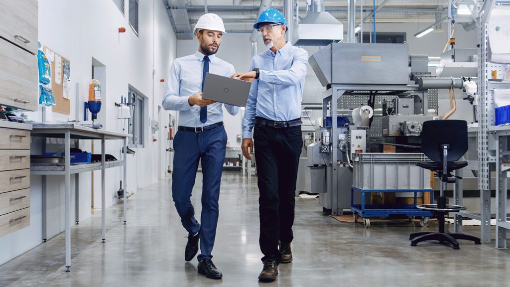 Two men in hard hats and business attire walk through a factory. One holds a laptop, pointing at the screen as they discuss something. The factory has various machines and equipment in the background, with a polished concrete floor and bright lighting.