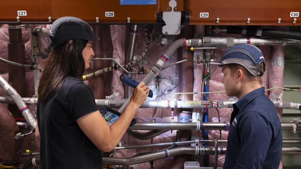Two individuals wearing caps and work uniforms are inspecting a complex system of pipes and machinery. One is using a flashlight and a tablet while pointing at the equipment; the other is observing attentively. The background consists of various metal components and insulation material.