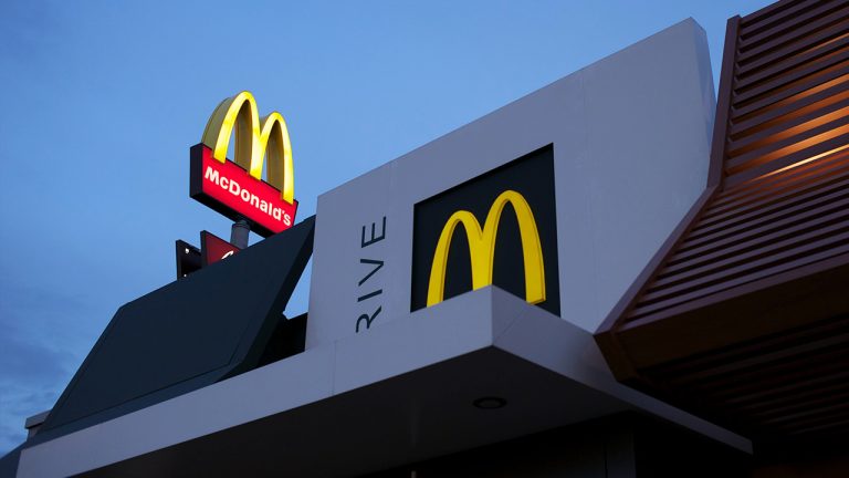 Exterior view of a McDonald's restaurant during evening. The iconic golden arches logo is prominently displayed on the building and a roadside sign, both illuminated against a dusky sky.