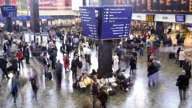 A busy train station with many people walking and standing. An overhead sign displays information about tickets, trains, and services. Some individuals are sitting on benches, and others are gathered around the sign. The atmosphere is bustling and dynamic.