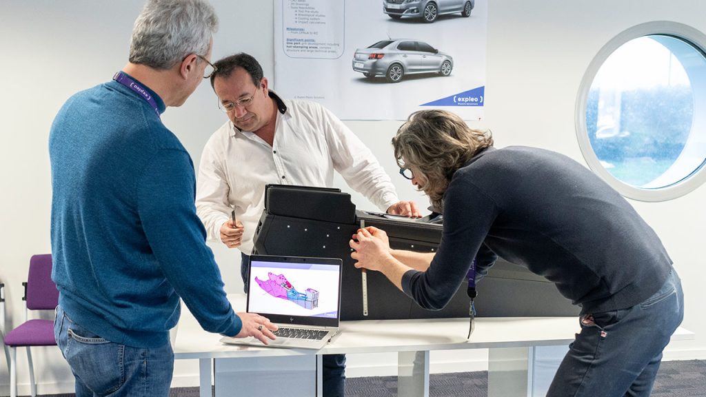 Three men are collaboratively working on a project, examining a black, box-like device on a table. One man is showing a diagram on a laptop. They're in a bright, modern room with posters of cars on the wall and a circular window.