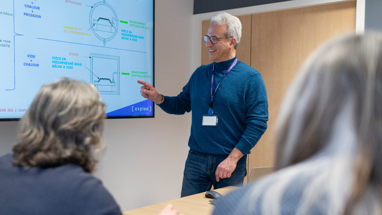 A man with gray hair and glasses is giving a presentation in a conference room, pointing to a diagram on a screen. Two people are seated at the table, observing. The presentation appears to involve technical or engineering concepts.