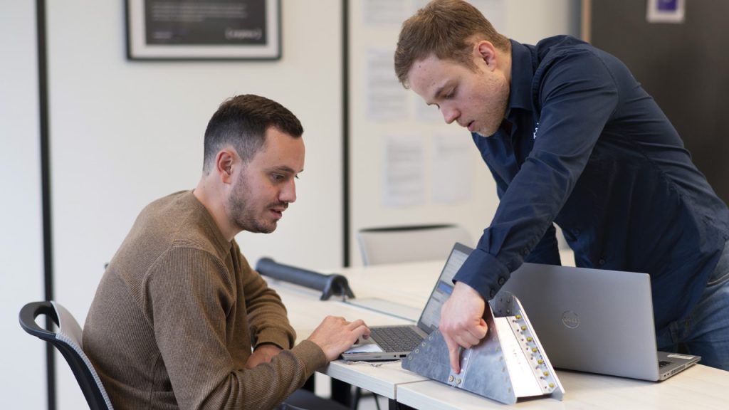 Two men working together in an office setting. One is seated and typing on a laptop, while the other, standing, is gesturing towards a metal device on the desk. Both appear focused and engaged in their task.