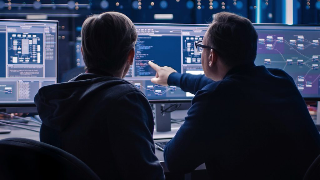 Two individuals are seated at a desk with multiple large computer monitors displaying code and data visualizations. One person is pointing towards the screen, appearing to explain something. The background is dark, with illuminated network equipment visible.