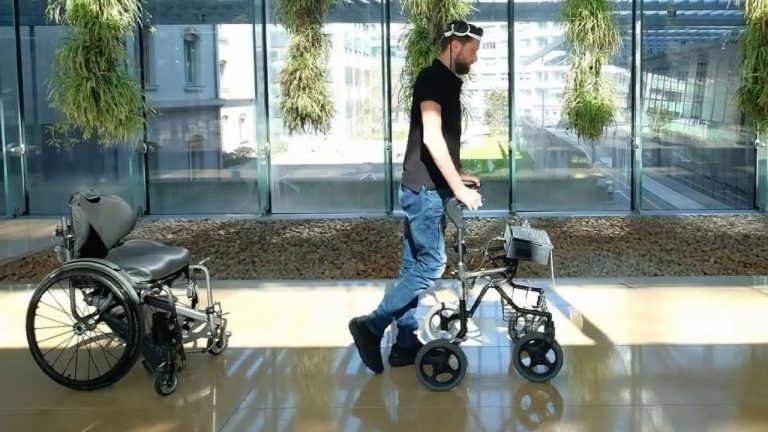 A man walks with the assistance of a walker through a sunlit corridor with large windows and hanging plants. He is wearing a cap, black t-shirt, and blue jeans. A wheelchair is visible to his left, indicating a transition from wheelchair to walker use.