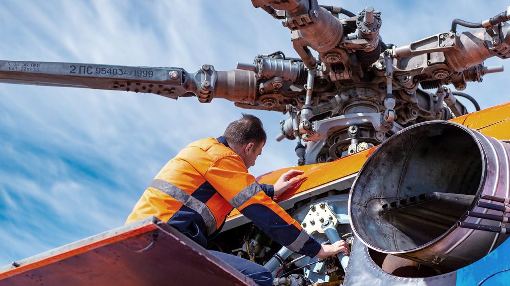 Worker in an orange and navy blue uniform performs maintenance on a helicopter's rotor system under a clear blue sky. The detailed mechanical components of the rotor assembly are visible.