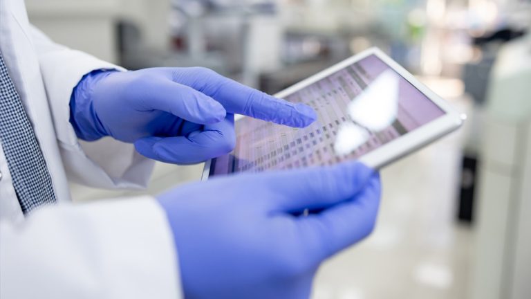 A person wearing a white lab coat and blue gloves holds a tablet. They are using one hand to touch the screen, which displays data in rows and columns. The background is blurred, suggesting an indoor, clinical or laboratory setting.