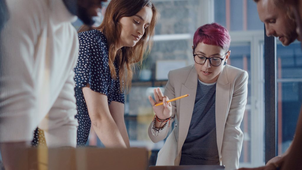 Four people are engaged in a collaborative discussion, standing around a table in a modern office environment. One of them, with short pink hair, is pointing at something on the table with a pencil. The atmosphere appears focused and cooperative.