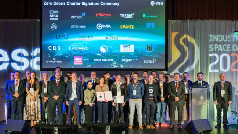 A group of people stands on a stage, posing for a photo in front of a screen displaying logos from various organizations. The screen reads "Zero Debris Charter Signature Ceremony". The event appears part of the "Industry Space Days 2023" hosted by ESA.
