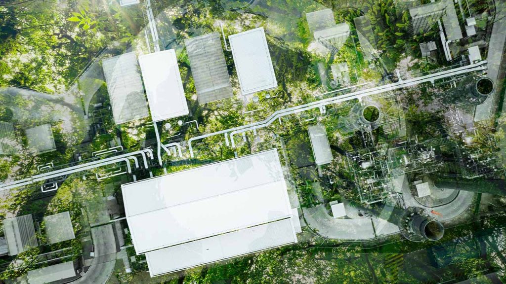 Aerial view of an industrial complex with multiple white-roofed buildings and surrounding greenery. Roads and pathways interconnect the structures, and trees and vegetation are visible throughout the area.