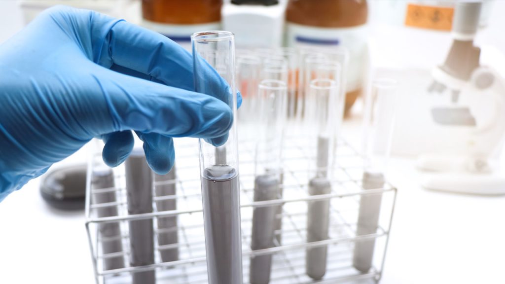 A gloved hand holding a test tube filled with a grey substance, placed in a rack among other similar tubes. In the background, there are brown lab bottles and a blurred-out microscope, indicating a laboratory setting.