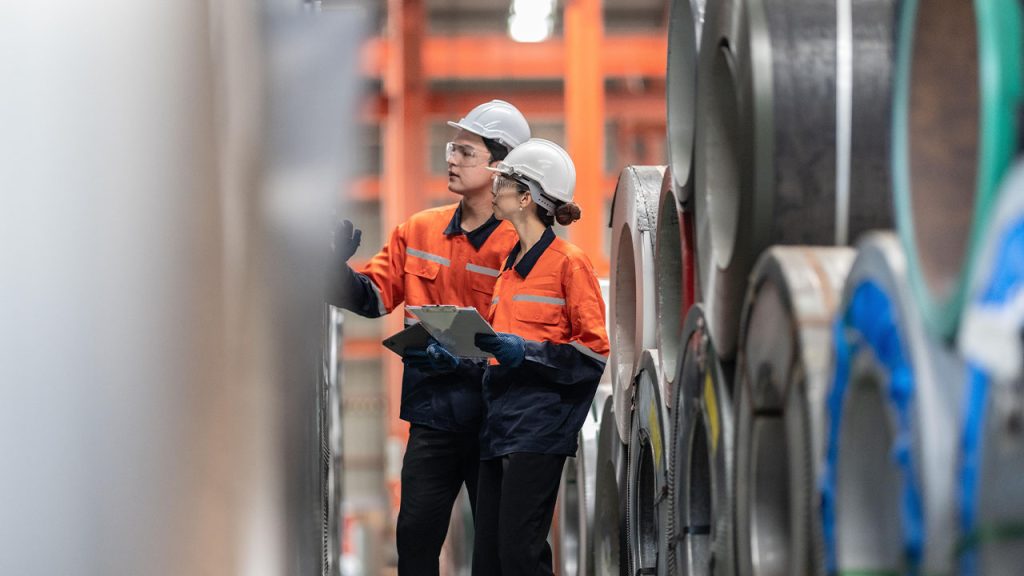 Two industrial workers wearing helmets and orange safety jackets inspect large metal rolls in a warehouse. One holds a clipboard, and they appear focused, amid a row of coiled metal sheets.