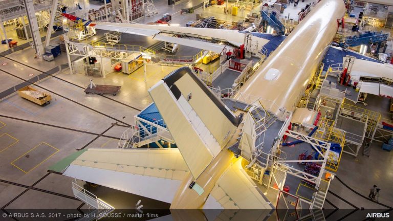An aerial view of an Airbus aircraft under assembly inside a large, bright factory, with workers, machinery, and scaffolding surrounding the plane. The plane’s yellowish body is partially constructed.