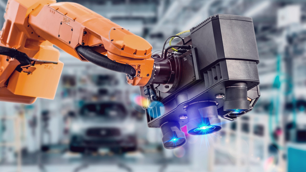 A robotic arm with a black camera or sensor attachment scans an object in a modern, blurred industrial or automotive factory setting. Blue lights on the device are illuminated.