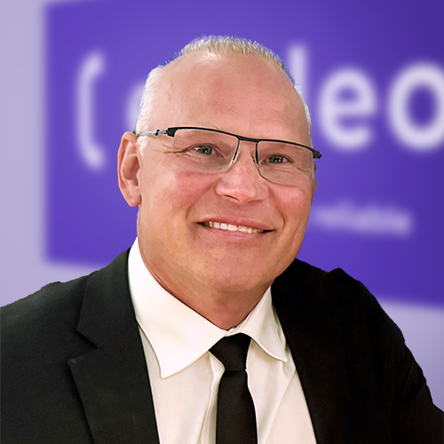 A smiling man with short blonde hair and glasses, wearing a black suit and tie, sits in front of a blurred purple background with partial white text.