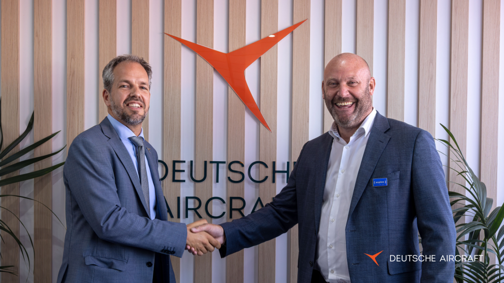 Two men in business suits shake hands and smile in front of a "Deutsche Aircraft" sign on a striped wooden wall, with the company's orange logo above them and green plants on either side.
