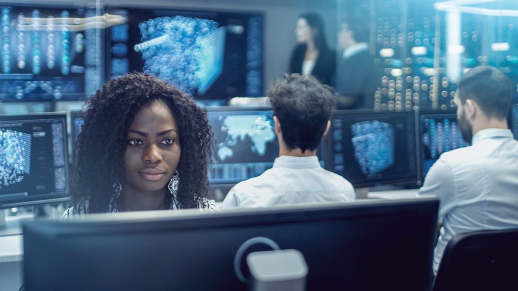 A woman sits at a computer in a modern office with multiple monitors displaying data visualizations. Two men work at nearby desks, and two people stand talking in the blurred background. The atmosphere is high-tech and collaborative.