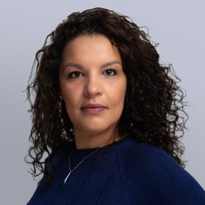 Woman with curly dark hair wearing a dark blue sweater and a necklace, looking confidently at the camera against a plain light gray background.