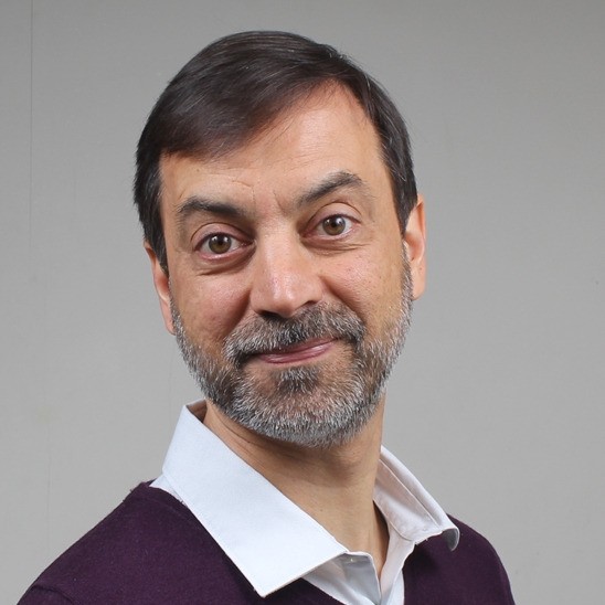 Un homme d'âge moyen aux cheveux bruns courts et à la barbe sourit gentiment à l'appareil photo. Il est vêtu d'une chemise blanche à col sous un pull foncé, et se tient devant un fond gris clair uni.