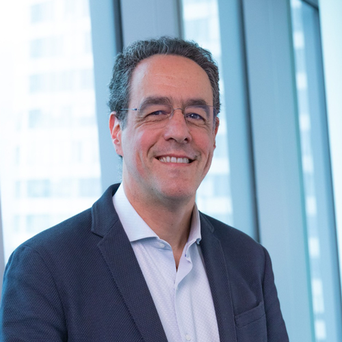 Un homme d'âge moyen aux cheveux bruns et courts, portant des lunettes et une veste de costume, sourit devant les hautes fenêtres d'un immeuble de bureaux moderne.