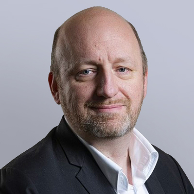 A middle-aged man with light skin, a beard, and thinning hair, wearing a dark suit jacket over a white shirt, poses against a plain light gray background.