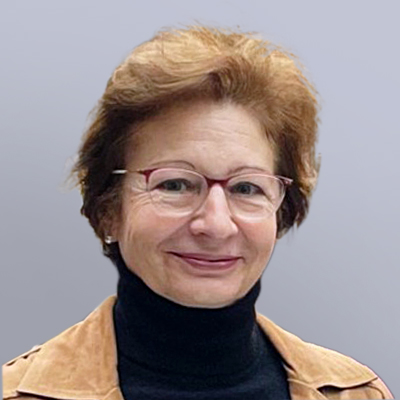 A woman with short light brown hair and glasses smiles at the camera. She is wearing a black turtleneck and a tan jacket, with a light gray background behind her.