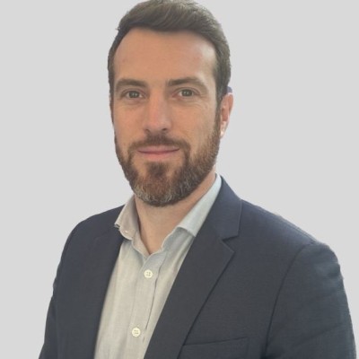 Un homme aux cheveux bruns courts et à la barbe, vêtu d'une veste de costume sombre et d'une chemise claire, pose sur un fond gris clair uni.